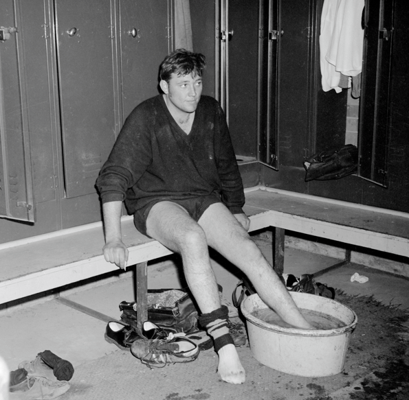 A vintage black and white photo of a rugby player in a locker room soaking his feet in a basin after a match, with boots and open lockers around him.